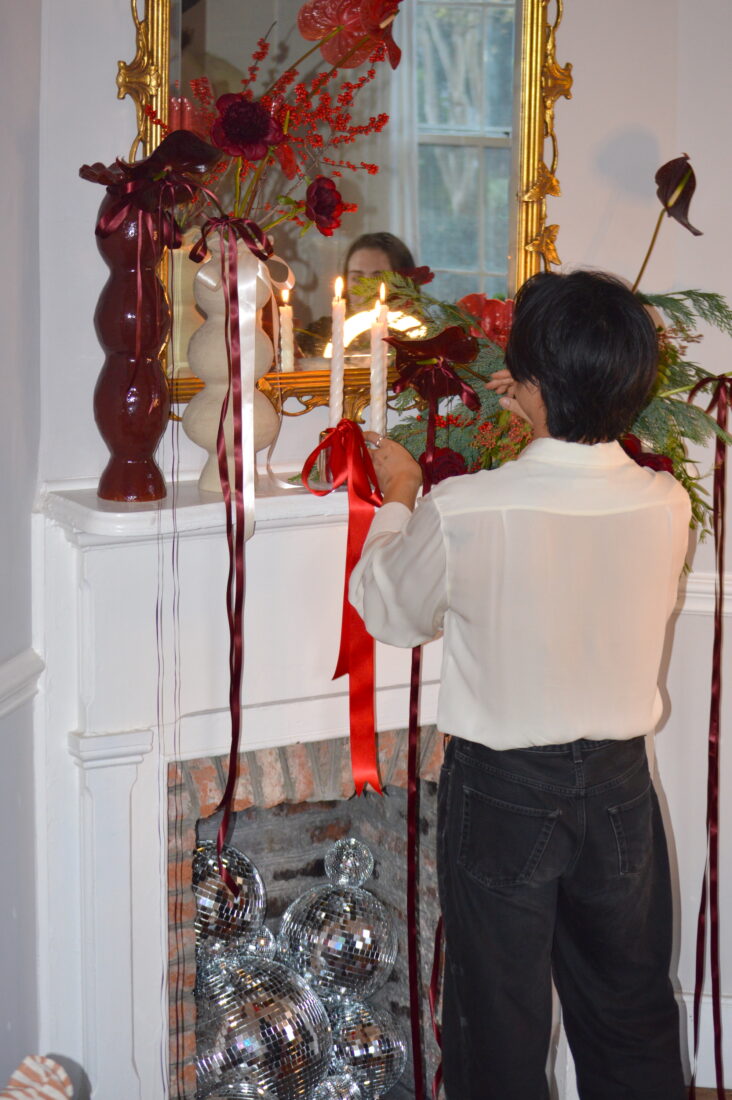 A person lights taper candles on a fireplace mantel with flowers and ribbon