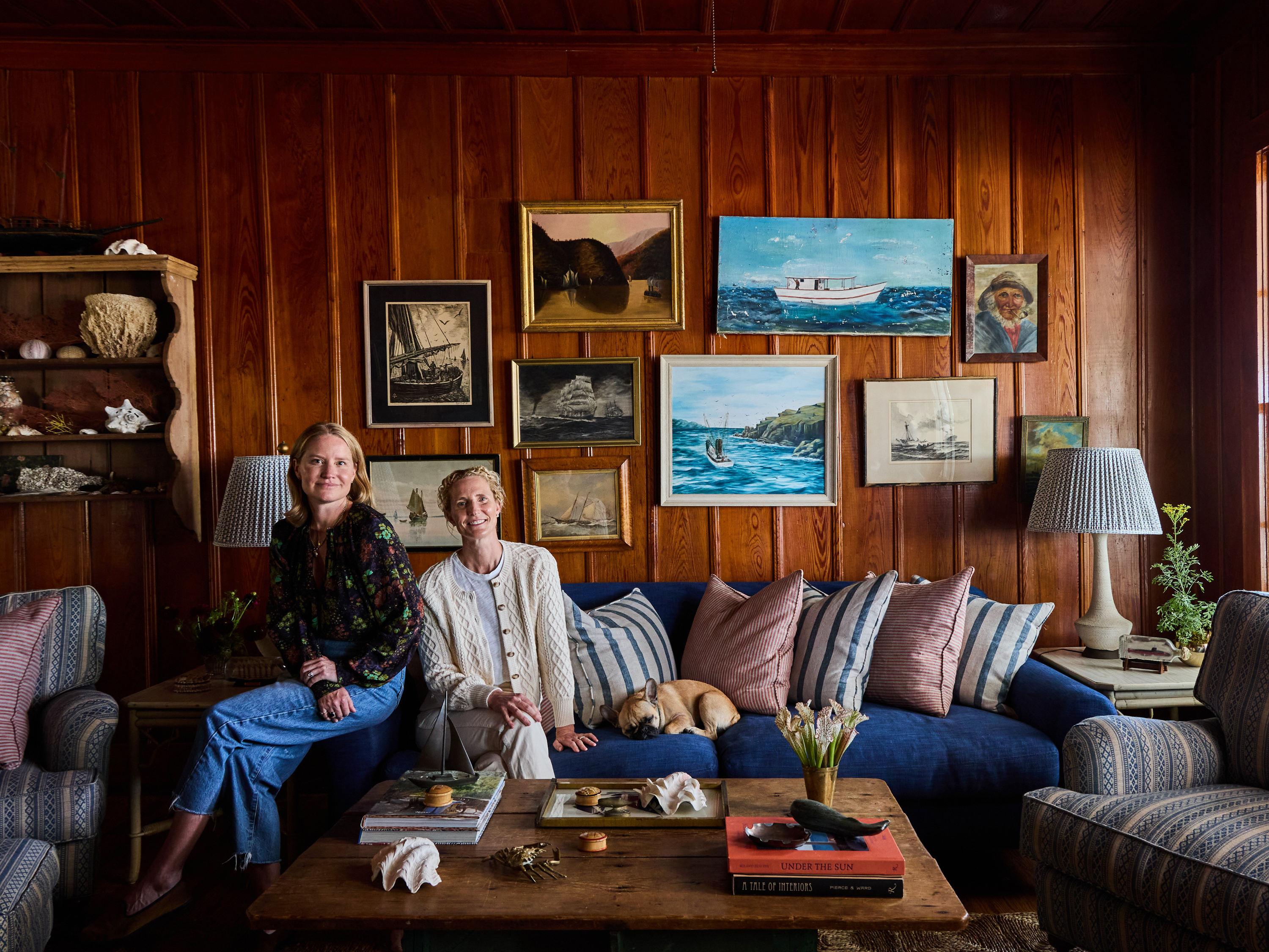 Two women and a dog sit on a couch in a wood paneled living room