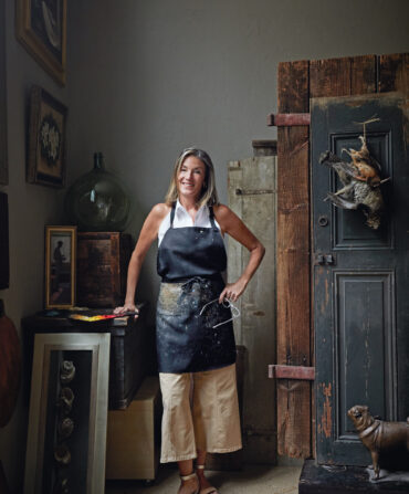 A woman with a black apron stands in an art studio with two birds hanging from the door