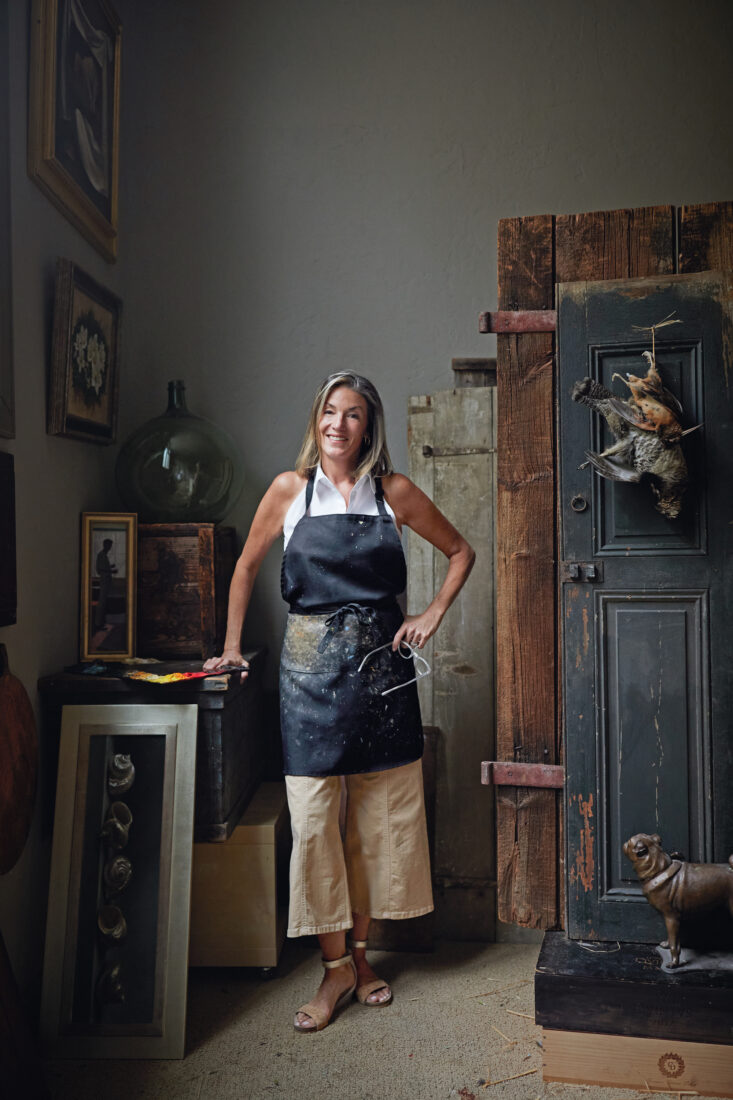 A woman with a black apron stands in an art studio with two birds hanging from the door