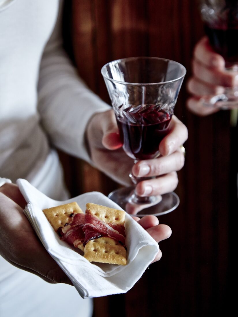 A woman holds a glass of red wine and a bacon wrapped cracker