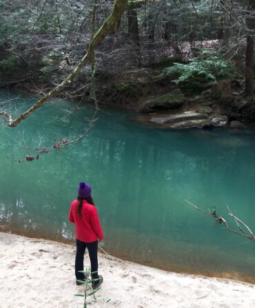 A person in a red coat stands by green water in a forest