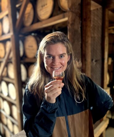 A woman stands in front of bourbon barrels. She holds a glass of bourbon