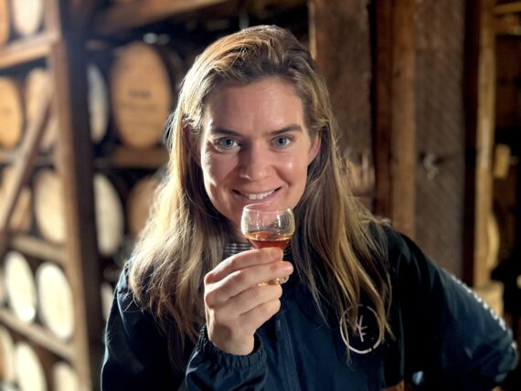 A woman stands in front of bourbon barrels. She holds a glass of bourbon