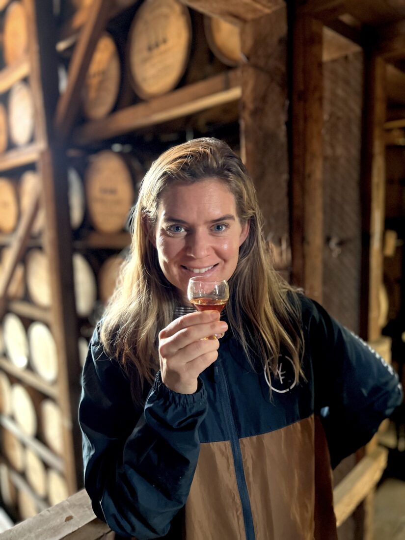 A woman stands in front of bourbon barrels. She holds a glass of bourbon