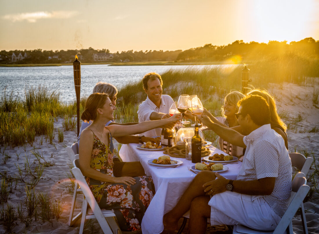 A group of people having dinner on the beach at a table lift their wine glasses to cheers