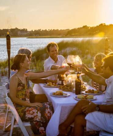 A group of people having dinner on the beach at a table lift their wine glasses to cheers