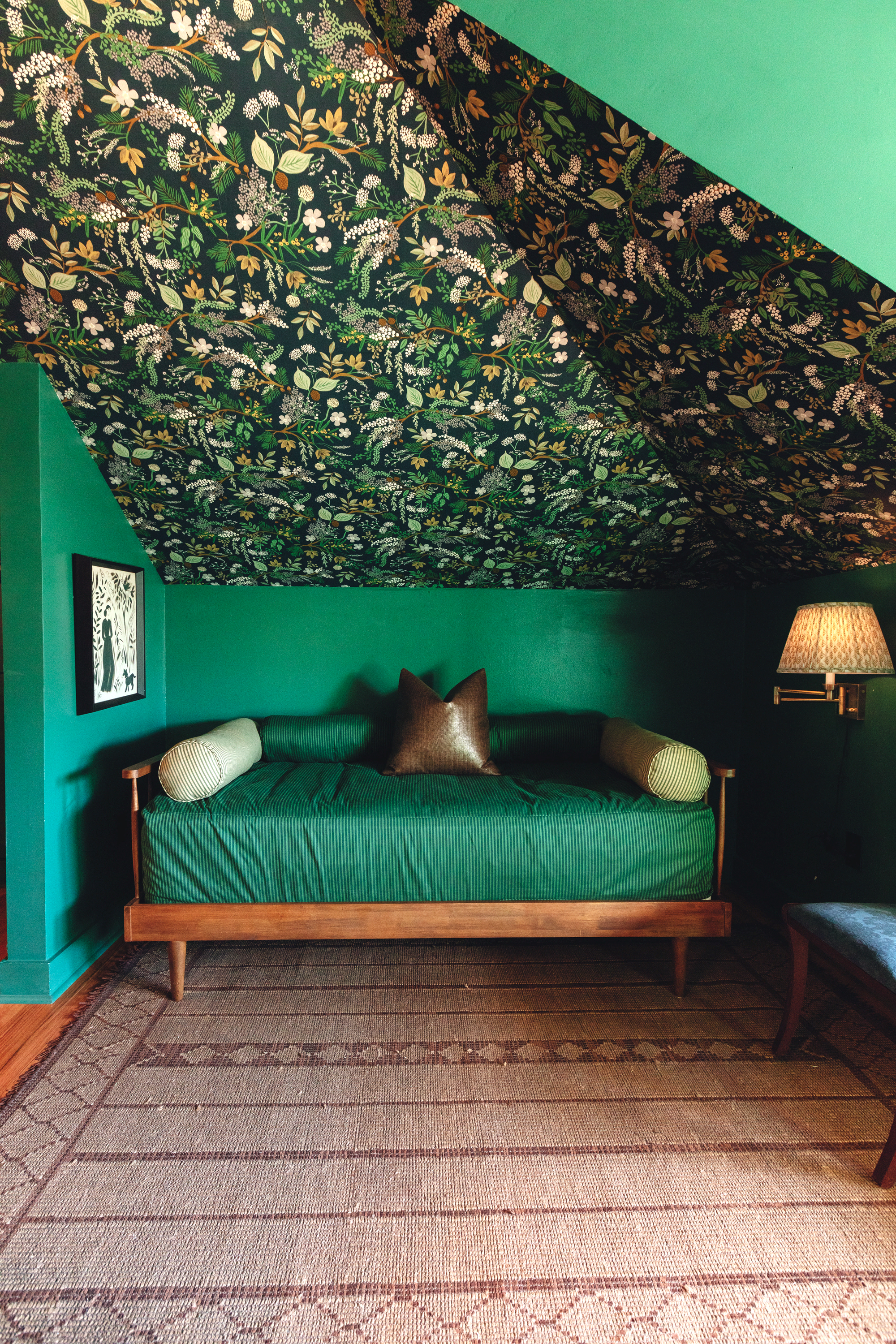 A corner of a guest room with a floral-printed pitched ceiling, green walls, and a green-cushioned and wood daybed