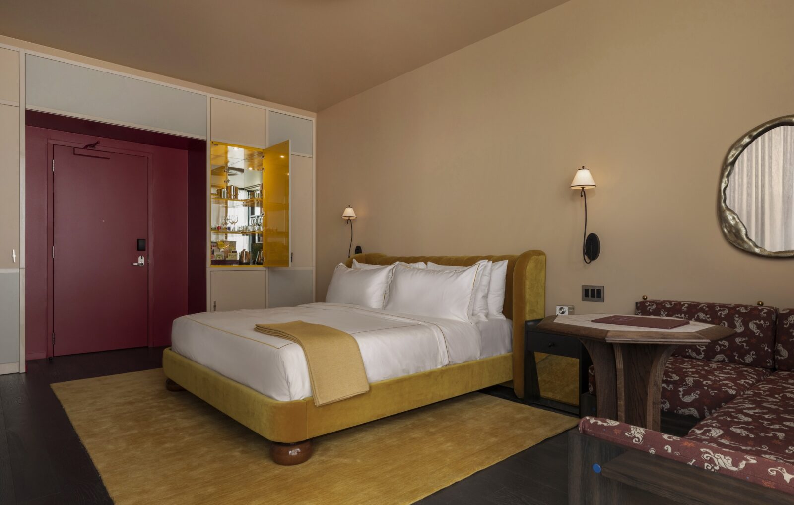 A cream guest room with yellow velvet bed and rug and a deep red door and cushioned banquette