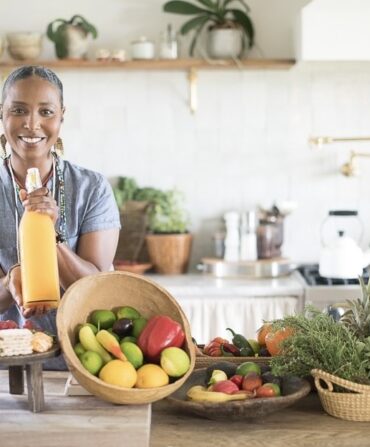 A woman in a kitchen holds up a bottle of juice. There are bowls of fruit around the counter