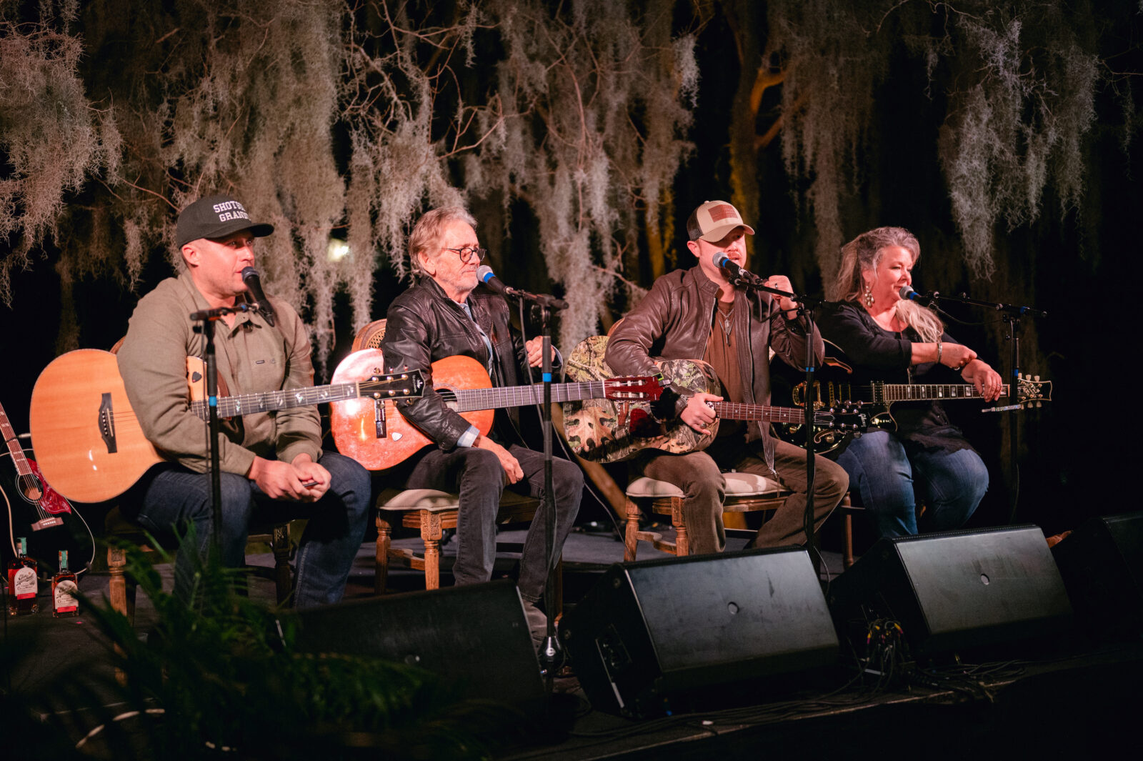 Four singers sit on a stage with guitars and microphones under branches with spanish moss