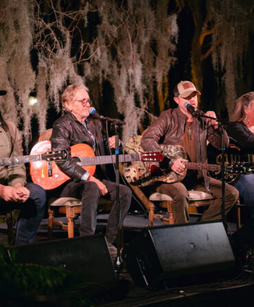 Four singers sit on a stage with guitars and microphones under branches with spanish moss