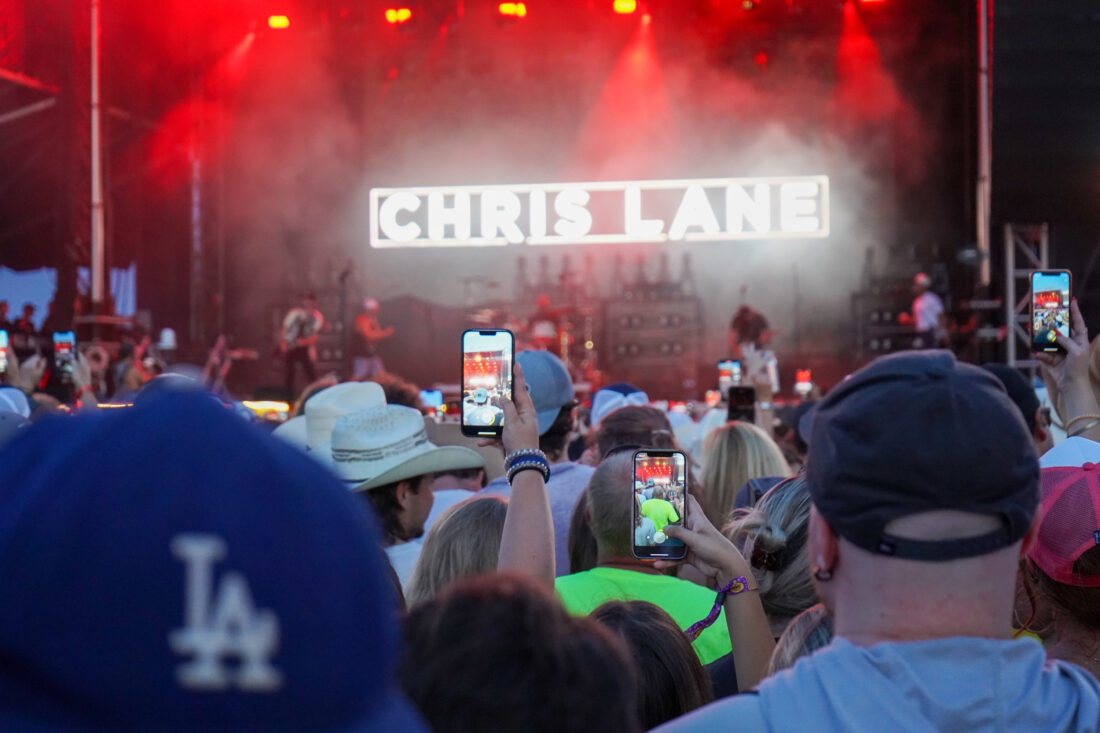 A crowd in front of a stage at a festival; people hold up phones to record the show