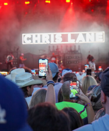 A crowd in front of a stage at a festival; people hold up phones to record the show