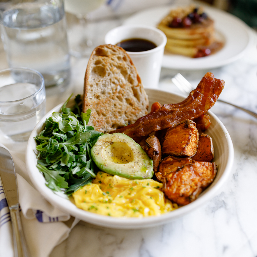 A bowl with two scrambled eggs, bacon, half avocado, dressed greens, roasted sweet potatoes, half slice of rustic sourdough