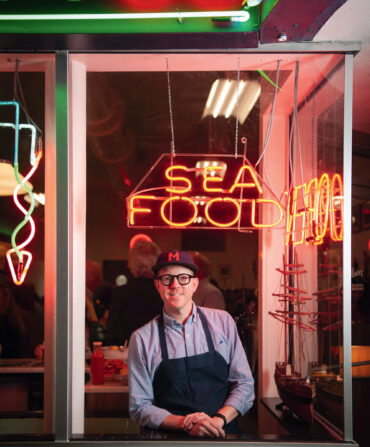 A chef stands in a restuarant window decorated with neon signs