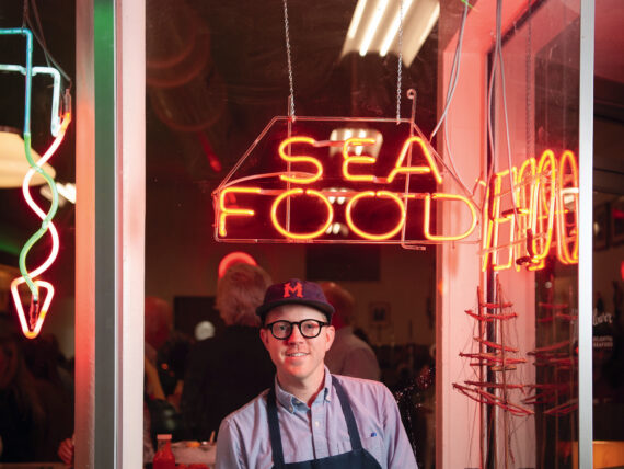 A chef stands in a restuarant window decorated with neon signs