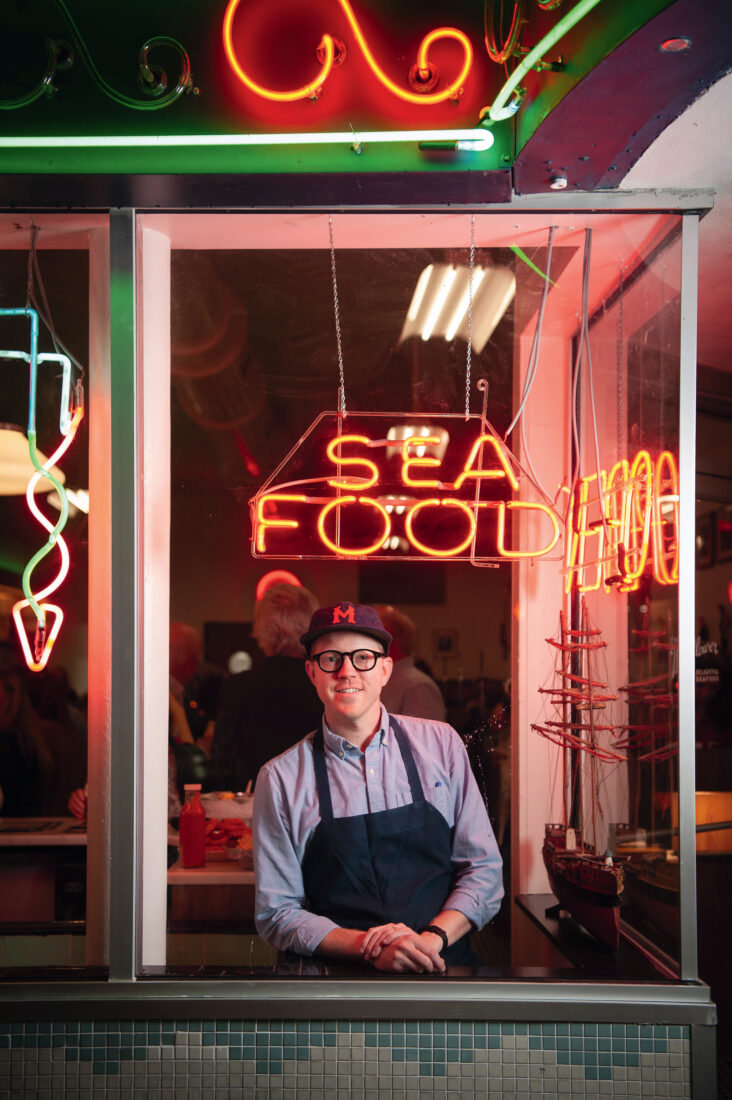 A chef stands in a restuarant window decorated with neon signs