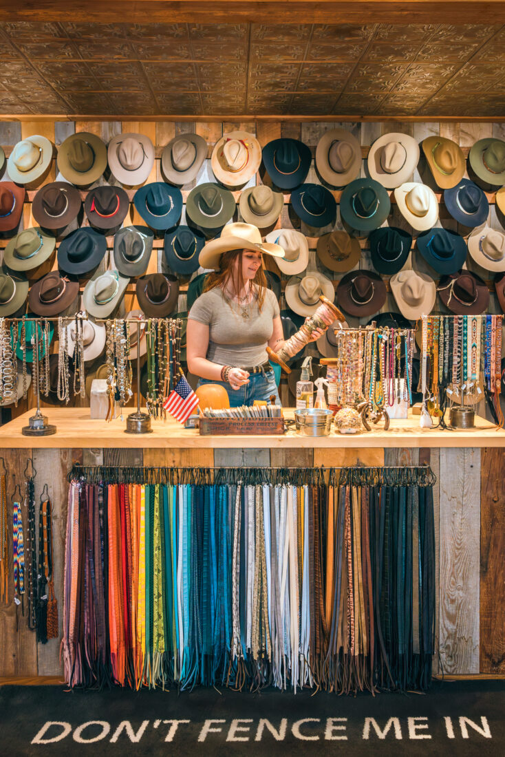 Inside a hat shop with colorful strips of leather, a wall of hats, and a woman behind a counter