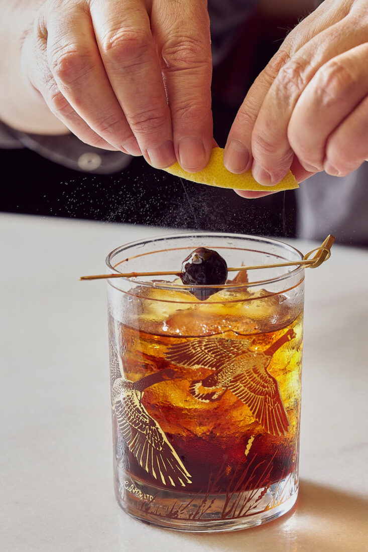 A hand twists a lemon peel over an old-fashioned