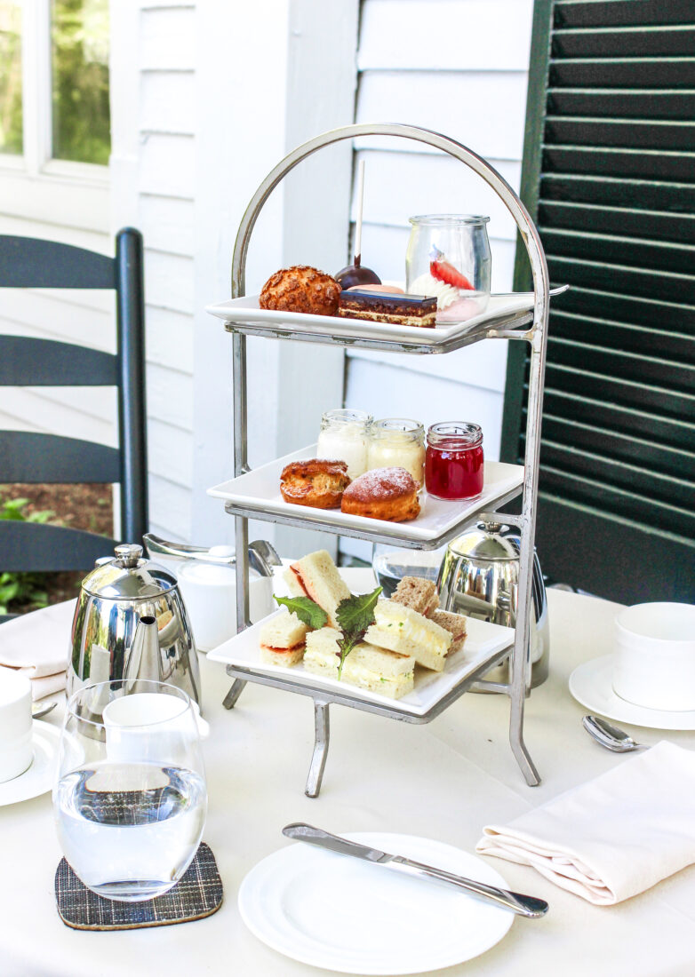 A tiered tray of tea snacks with a pot of tea on a porch table