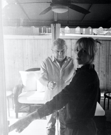 A portrait in black and white of a woman and older man. The woman stands in a doorway with the man behind her.