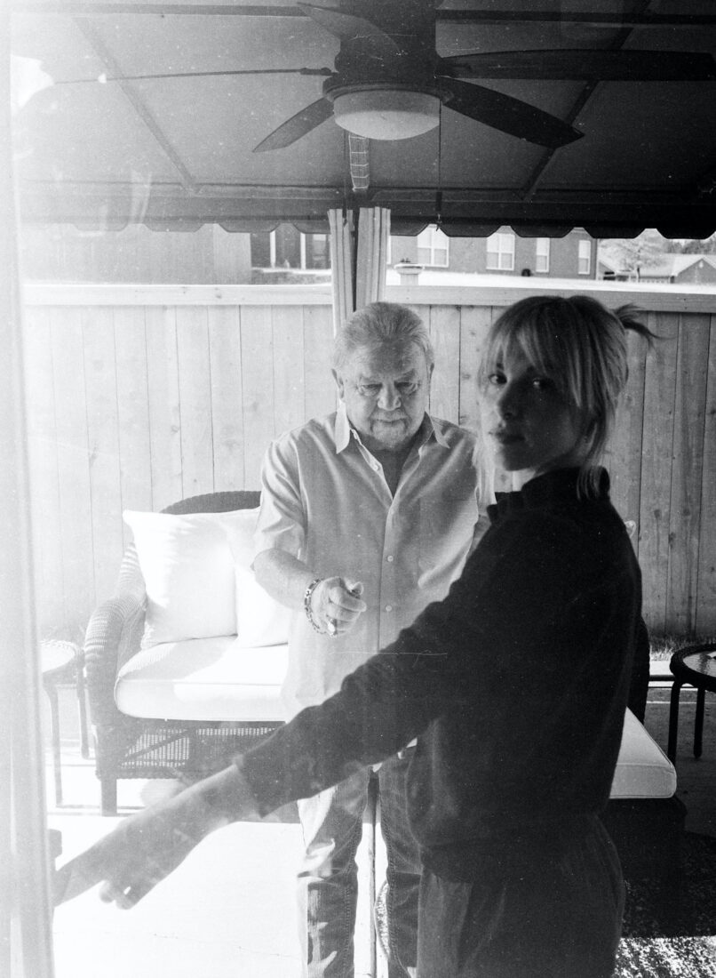 A portrait in black and white of a woman and older man. The woman stands in a doorway with the man behind her.