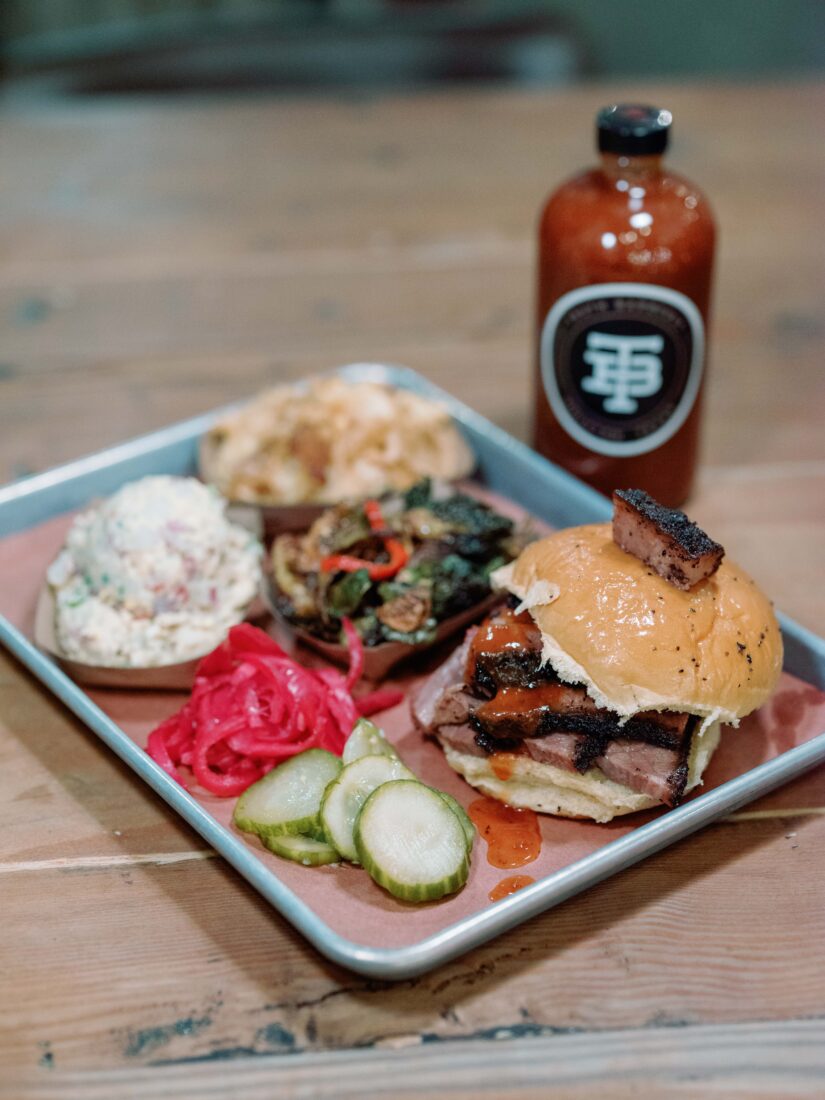 A sliced brisket sandwich with potato salad, tater tot casserole, and brussels sprouts on the side.
