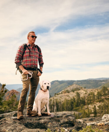 A man and dog stand on a rock against a mountain landscape