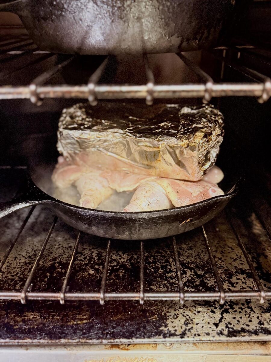 A flattened chicken under a tinfoiled wrapped brick in a cast iron pan in an oven