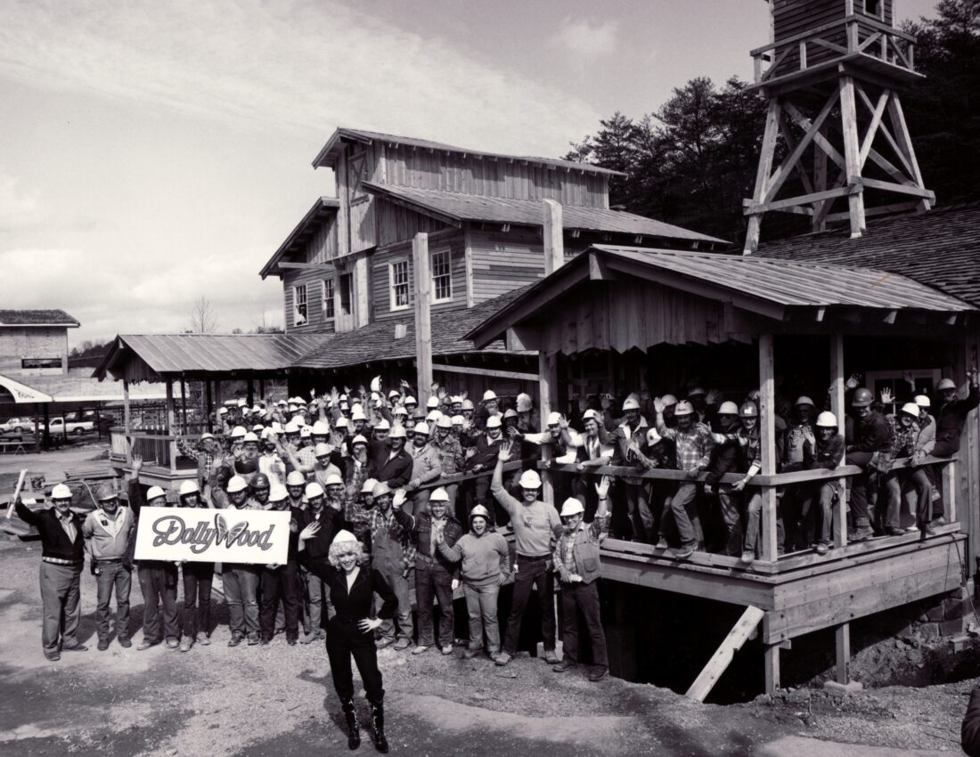 Dolly Parton and construction workers smile and wave at a camera at dollywood