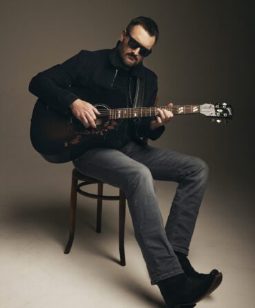 A portrait of a man holding a guitar and sitting on a stool