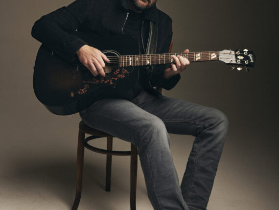 A portrait of a man holding a guitar and sitting on a stool