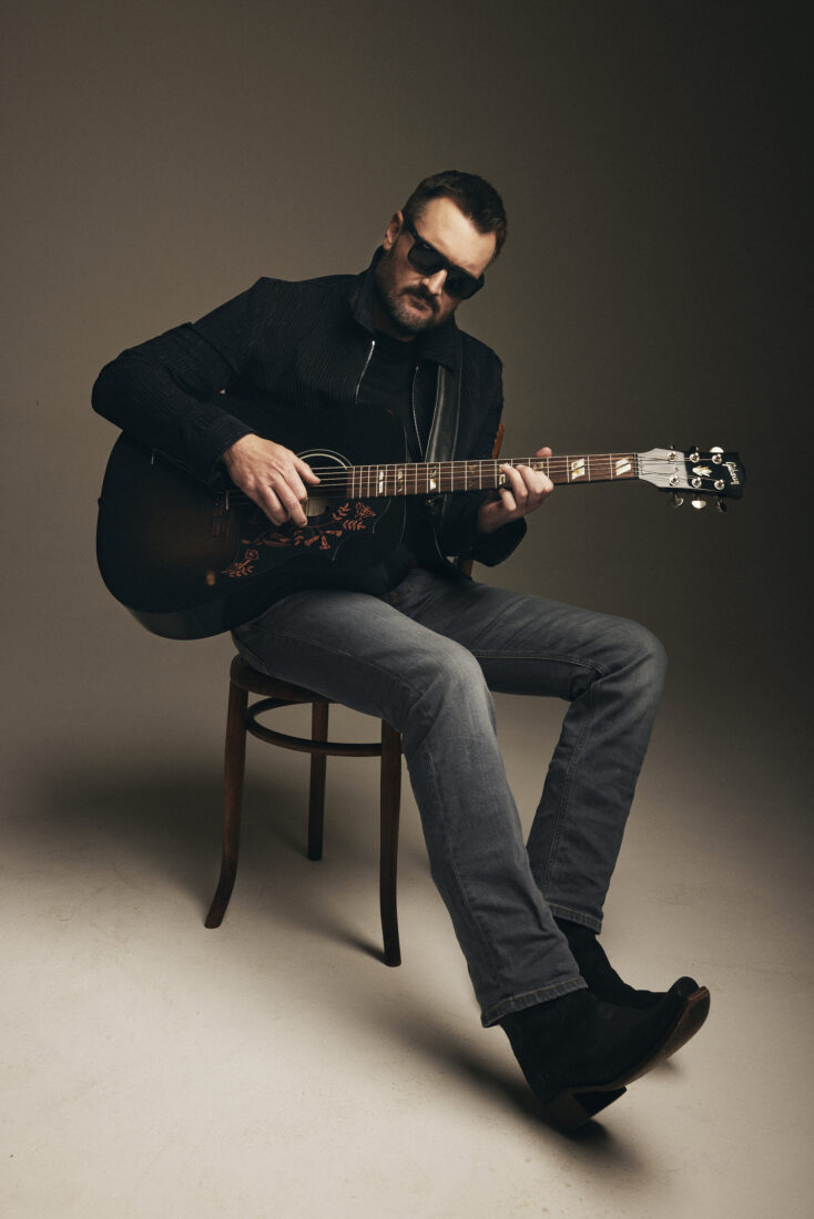 A portrait of a man holding a guitar and sitting on a stool