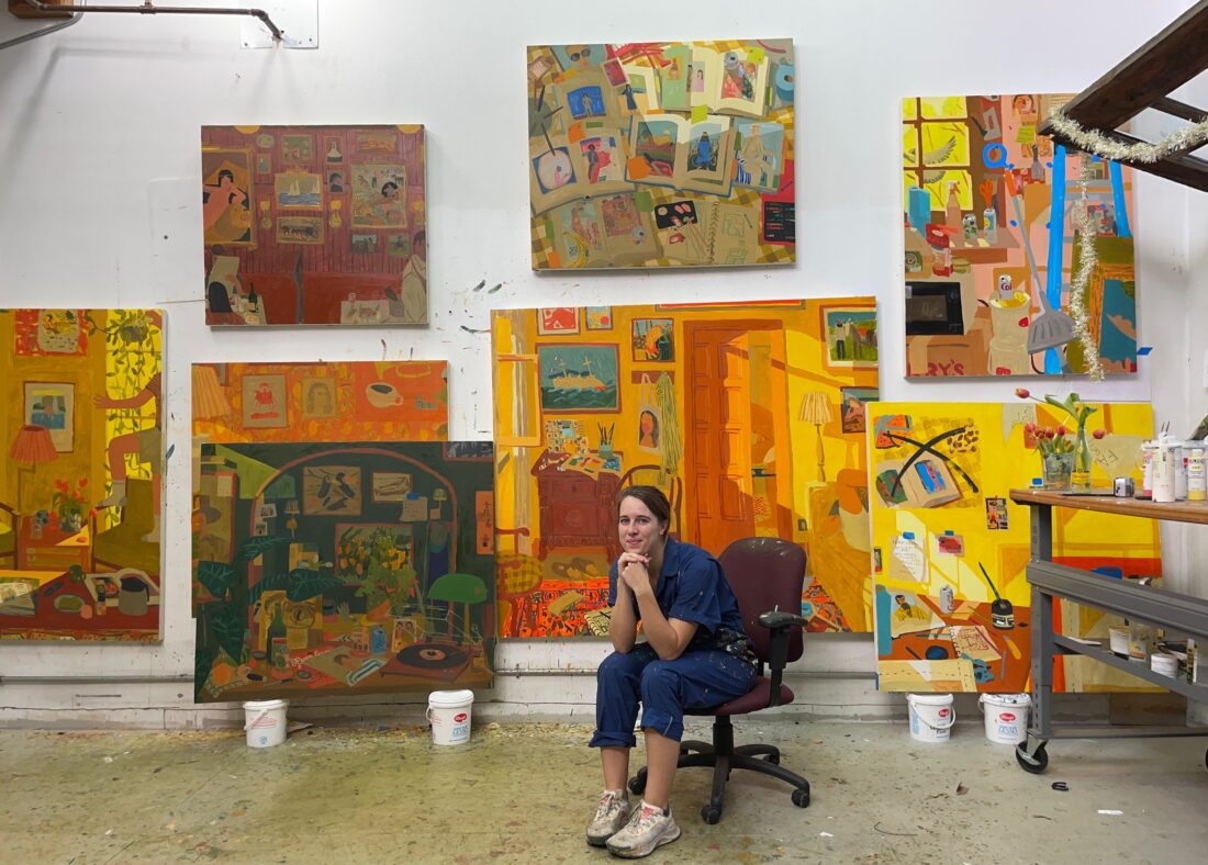 A woman sits in an art studio surrounded by colorful large paintings