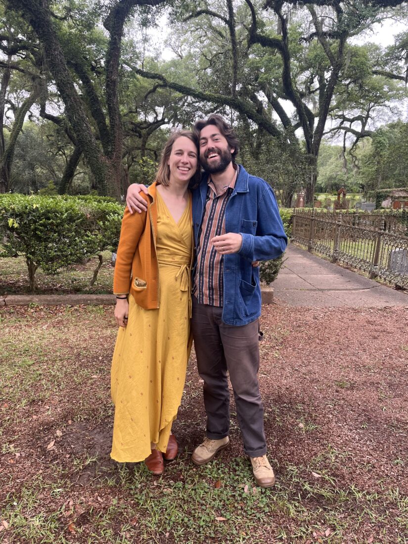 A woman in a yellow dress and man in a denim jacket smile in a graveyard