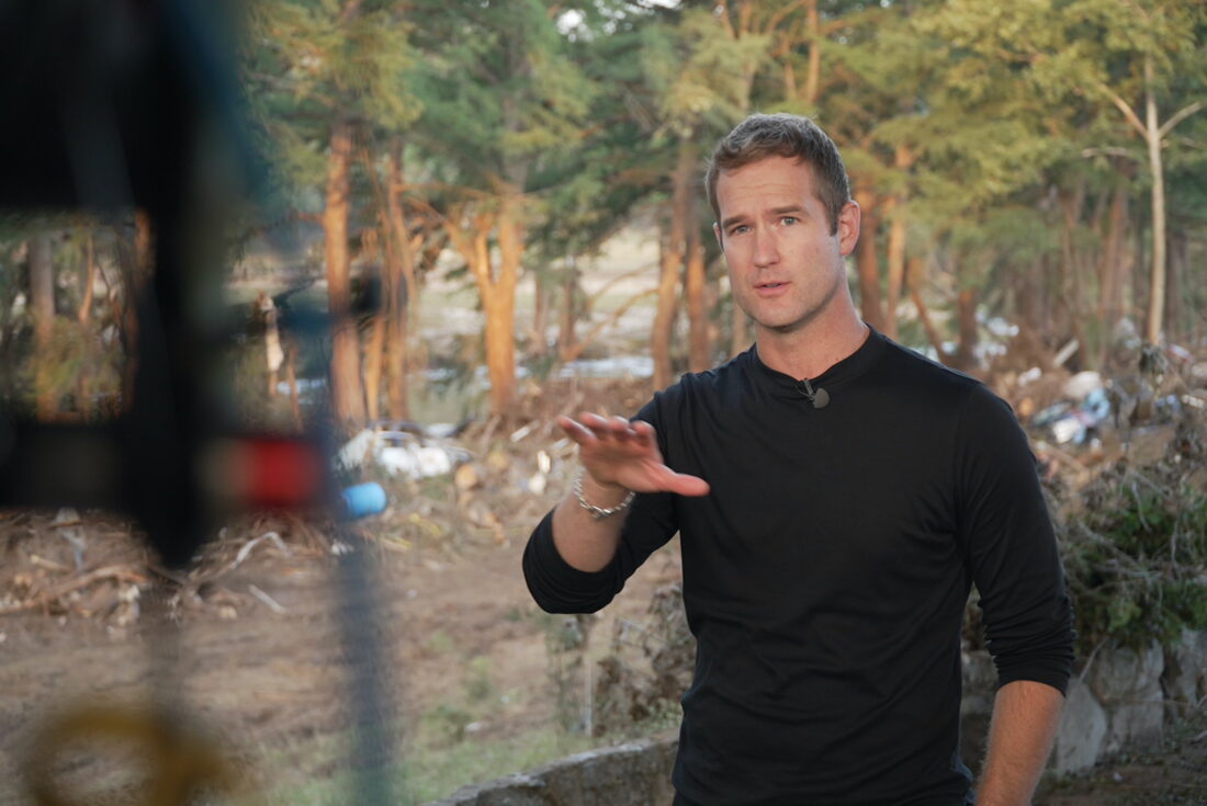 A man stands in front of flood wreckage, talking to a camera