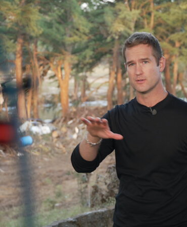 A man stands in front of flood wreckage, talking to a camera