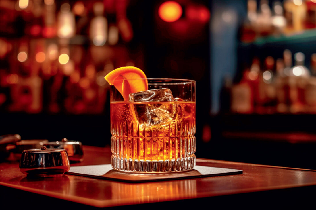 A glass of bourbon with an orange garnish on a table in a dimly lit bar