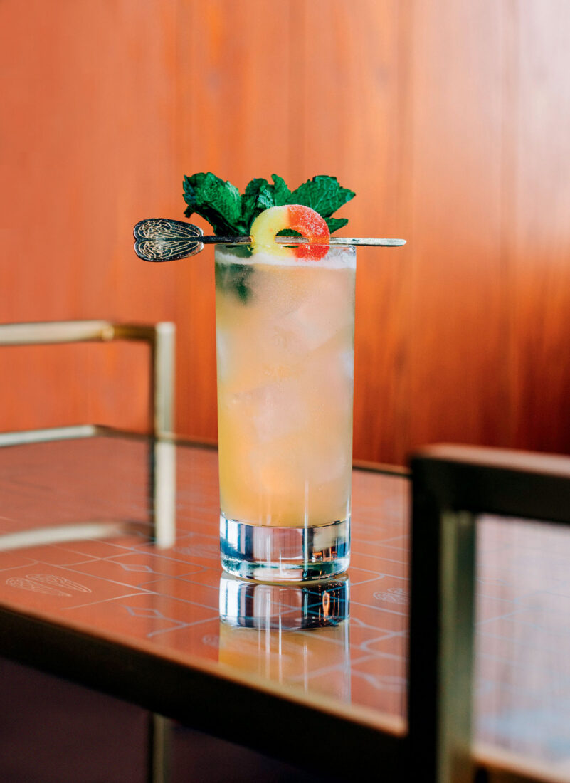 A tall cocktail glass with a mint garnish and peach ring cady on a skewer