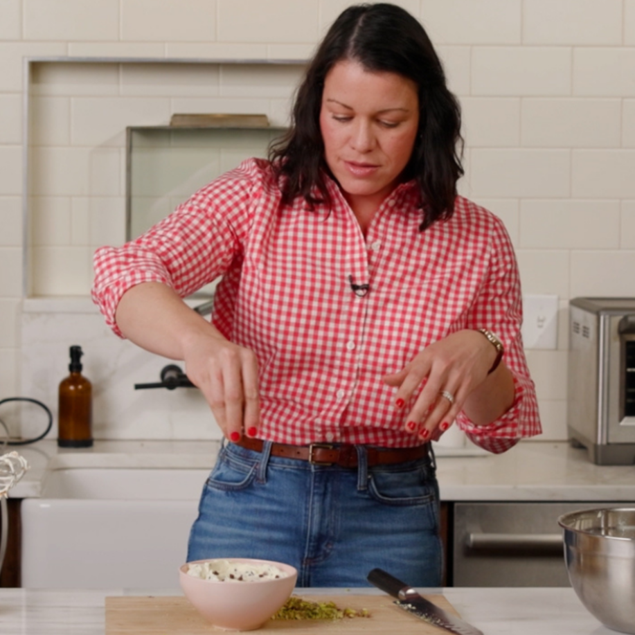 a woman in a red gingham shirt and jeans sprinkles chocolate chips into a cannoli dip