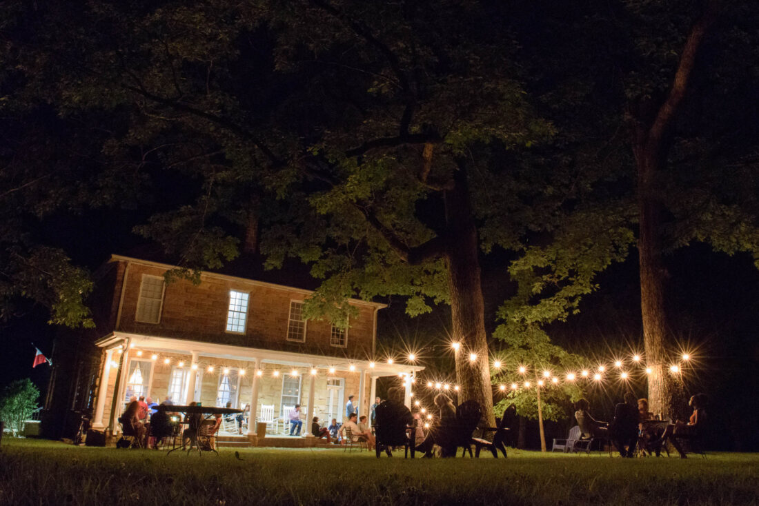 A group of people sit under string lights in a backyard