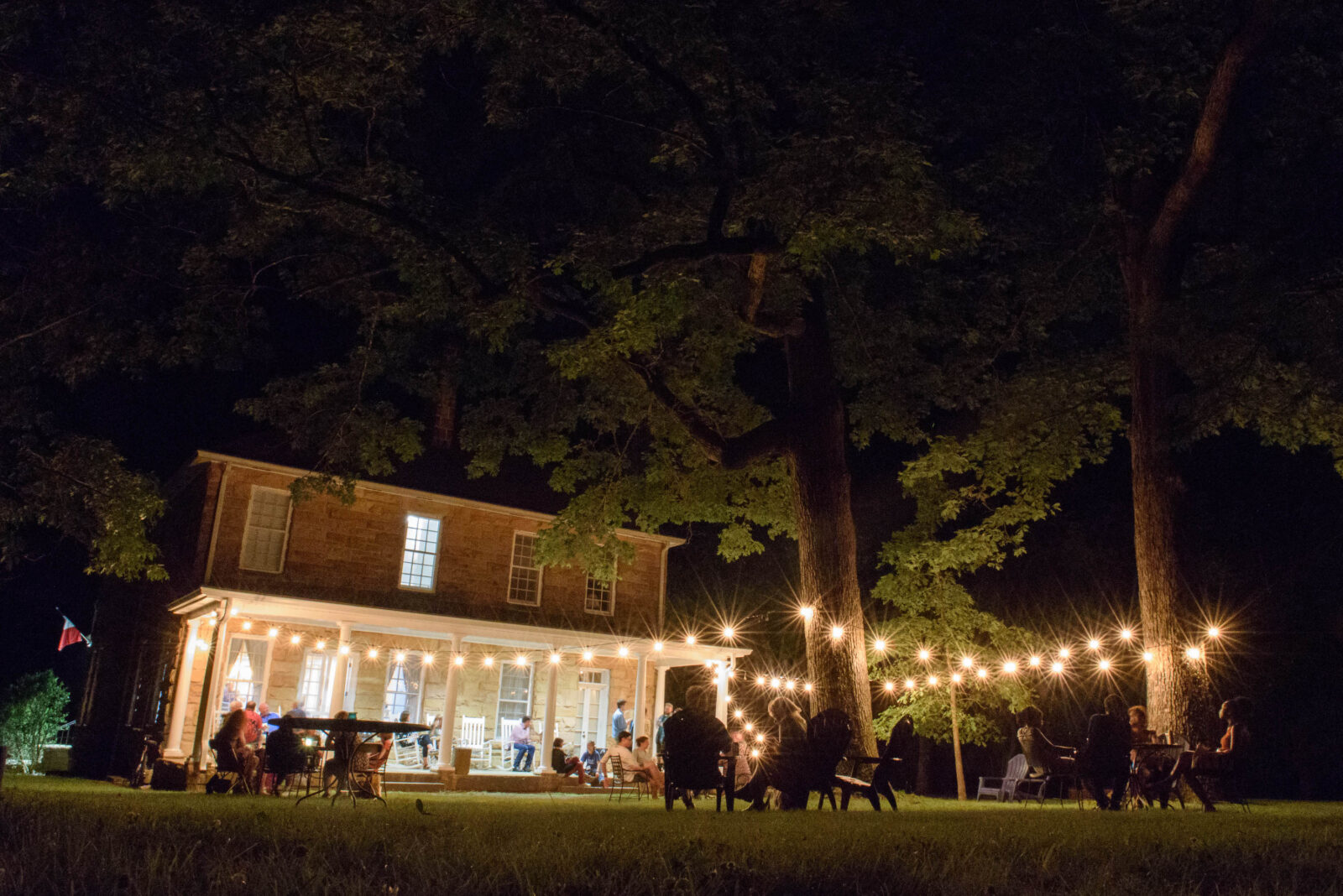 A group of people sit under string lights in a backyard