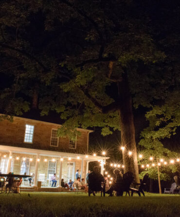 A group of people sit under string lights in a backyard