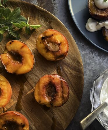 Grilled peaches on a wooden platter with a Bourbon and Brown-Sugar Butter Glaze, whipped cream, and baby basil leaves