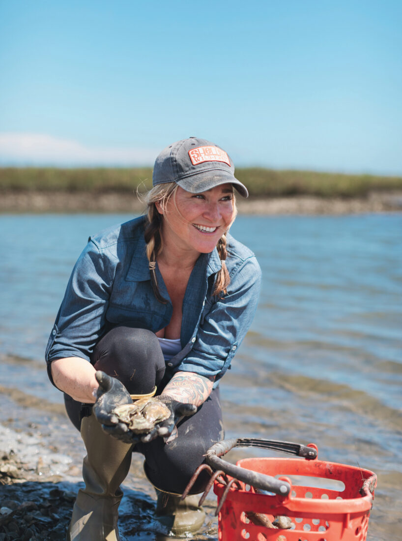 A woman harvests oysters from a marsh and smiles next to a red basket