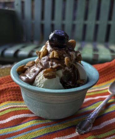 Ice cream sundae with bourbon-hot fudge, a bourbon-soaked cherry, and toasted pecans in a rustic mountain setting with a colorful striped napkin and spoon. Rusted old metal glider in background