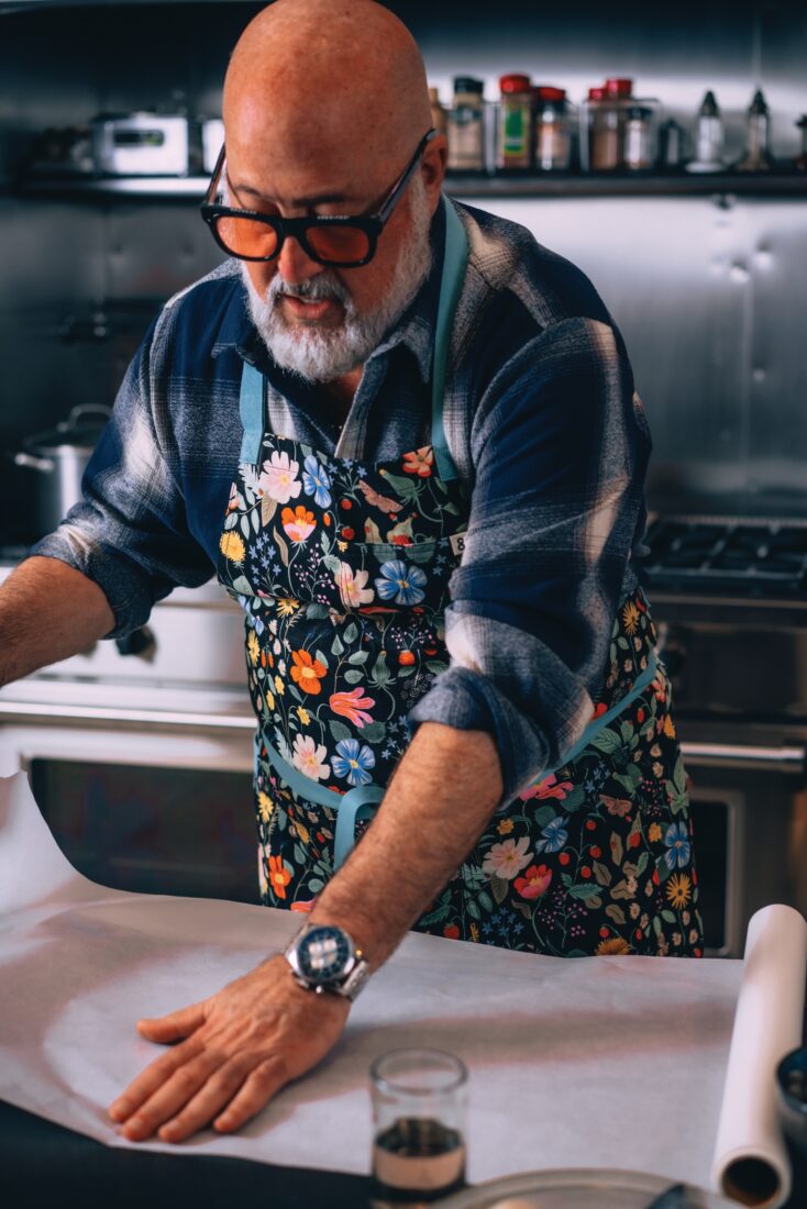 A chef in a floral apron rolls out parchment paper in a kitchen