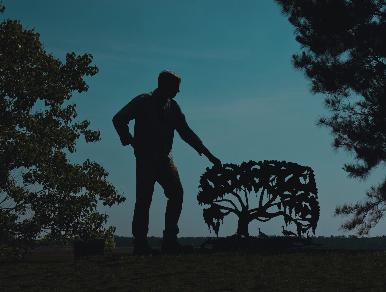 A man stands at dusk with a metal sculpture of a cut-out tree