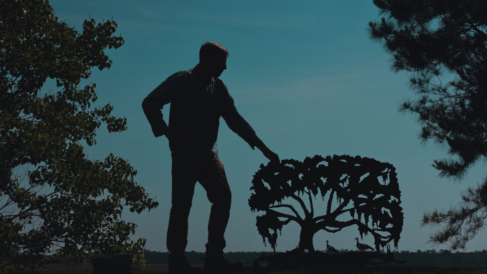 A man stands at dusk with a metal sculpture of a cut-out tree